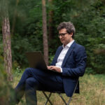 Man working on laptop in a green forest, blending freelance work, work at home flexibility, vacation, relaxation, and healthy work life balance outdoors.