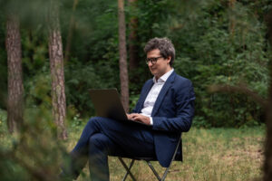 Man working on laptop in a green forest, blending freelance work, work at home flexibility, vacation, relaxation, and healthy work life balance outdoors.